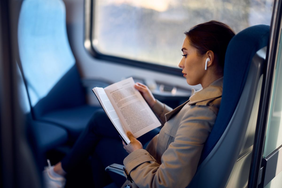 Young,Female,Passenger,Listening,Music,On,Earbuds,And,Reading,Book